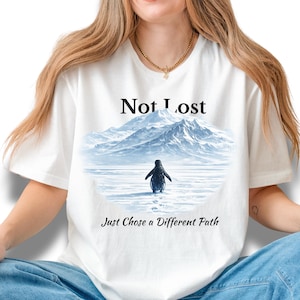 May include: White crew neck t-shirt with a penguin graphic walking on ice, set against a mountain scene. The text reads "Not Lost, Just Chose a Different Path". The shirt is a casual wear item.