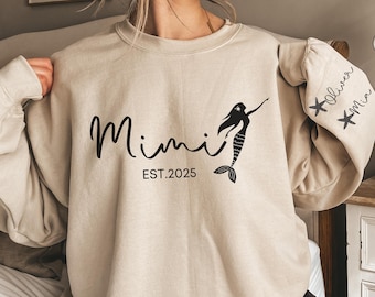 May include: A beige sweatshirt featuring the word "Mimi" in black script, a mermaid design, and "EST.2025". The right sleeve displays the names "Oliver" and "Mia" in black. A relaxed, comfortable garment.