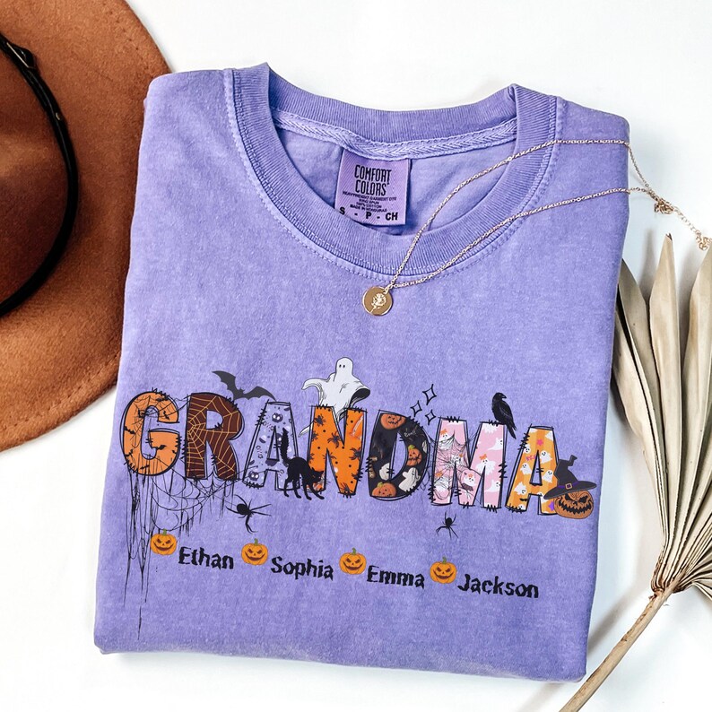 May include: A light purple Comfort Colors t-shirt with the word "GRANDMA" in a Halloween-themed design. The design includes bats, spiders, pumpkins, and the names Ethan, Sophia, Emma, and Jackson. A gold necklace is draped across the shirt.