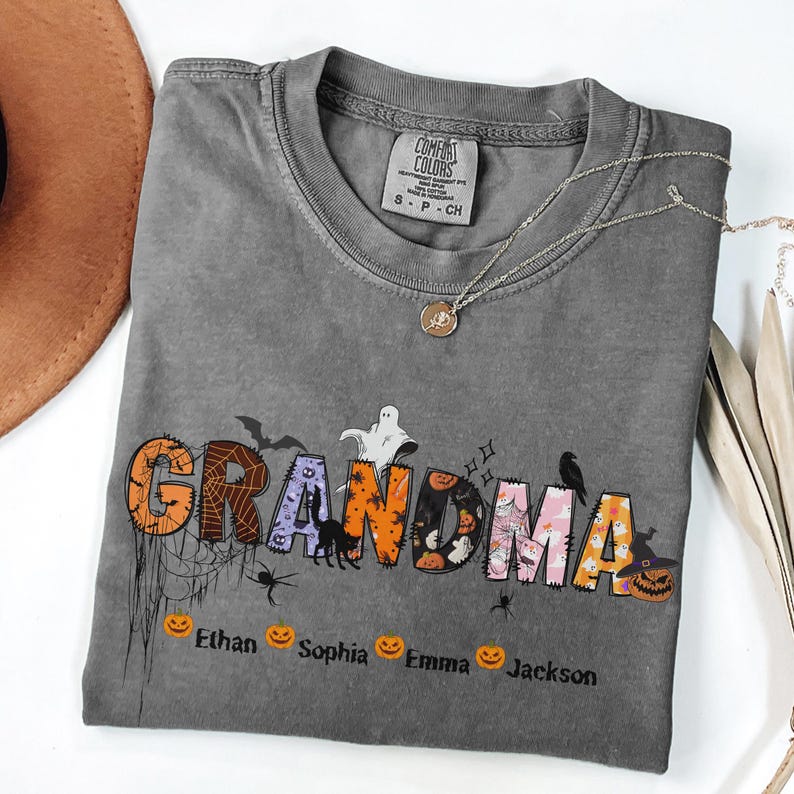 May include: A gray Comfort Colors t-shirt with the word "GRANDMA" in a Halloween-themed design. The letters are decorated with bats, spiders, pumpkins, and ghosts. Below the word are the names Ethan, Sophia, Emma, and Jackson.