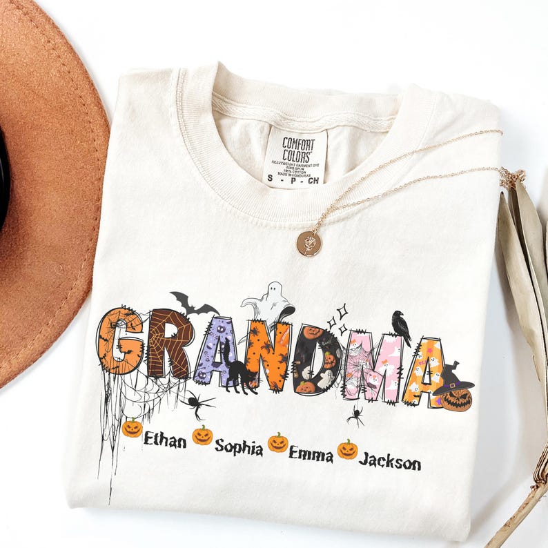 May include: Cream-colored Comfort Colors t-shirt with the word "GRANDMA" in a Halloween-themed design. The letters are decorated with bats, spiders, pumpkins, and ghosts. Names of grandchildren are below the word. A gold necklace and a brown hat are also in the image.