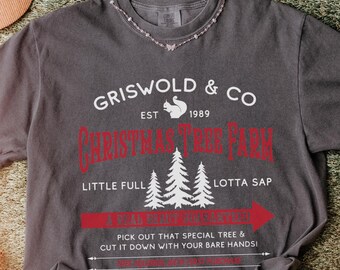 May include: Dark gray t-shirt with a red and white graphic that reads "Griswold & Co Christmas Tree Farm" with a squirrel logo and Christmas trees. The shirt also has the text "Pick out that special tree & cut it down with your bare hands!"