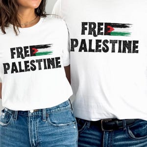 Może przedstawiać: Białe koszulki z napisem "FREE PALESTINE" czarnymi literami. Słowo "FREE" ma poziomy wzór pociągnięcia pędzlem w kolorach flagi Palestyny: czerwonym, białym, zielonym i czarnym. Koszulki są zestawione z niebieskimi dżinsami.