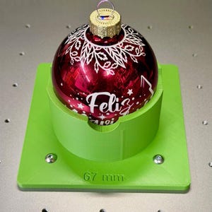 Op de afbeelding: Een rode kerstbal met witte bloemmotieven en het woord "Feliz" wordt op een groene standaard gepresenteerd. De bal heeft een gouden dop en heeft een diameter van 67 mm.