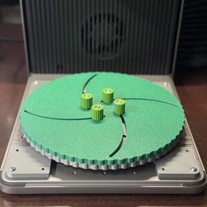 Puede incluir: Un plato giratorio verde con cinco pequeños botones verdes en la parte superior. El plato giratorio está sobre una base plateada.