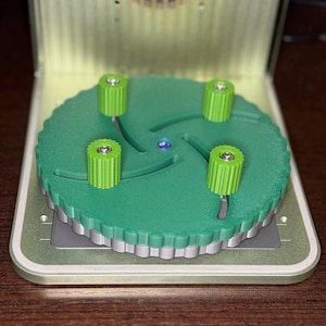 Puede incluir: Un plato giratorio verde con cuatro botones verdes. El plato giratorio está sobre una base gris.