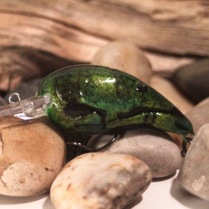 May include: Green and black fishing lure with a realistic crawfish design. The lure has a clear plastic bill and a black hook.