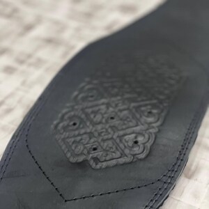 May include: A black leather belt with an embossed decorative design. The belt has a detailed pattern in the centre and is stitched along the edges. The design is symmetrical and appears to be a floral or geometric motif.