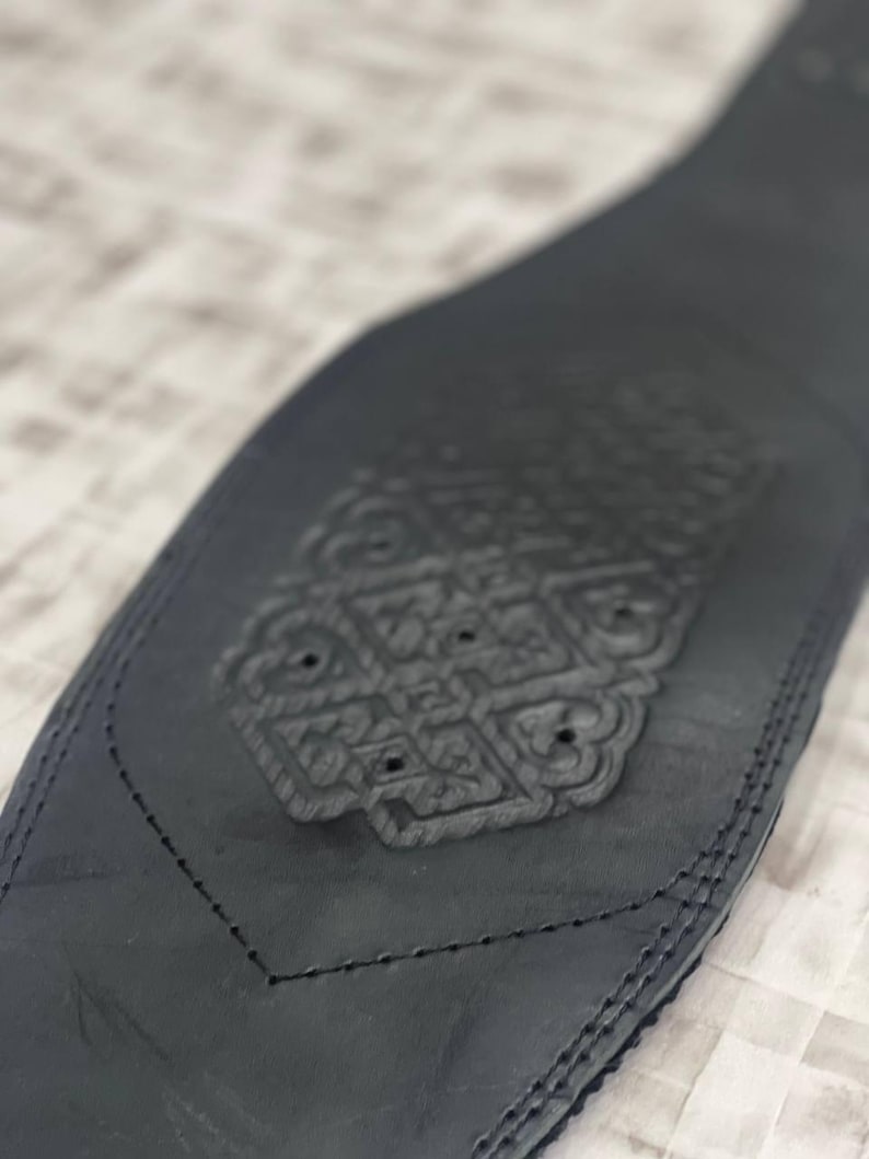 May include: A black leather belt with an embossed decorative design. The belt has a detailed pattern in the centre and is stitched along the edges. The design is symmetrical and appears to be a floral or geometric motif.