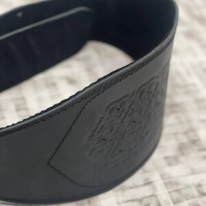May include: A close-up of a black leather belt with an embossed design. The belt is curved, showing the interior lining and stitched edges. The embossed pattern is visible on the front of the belt.