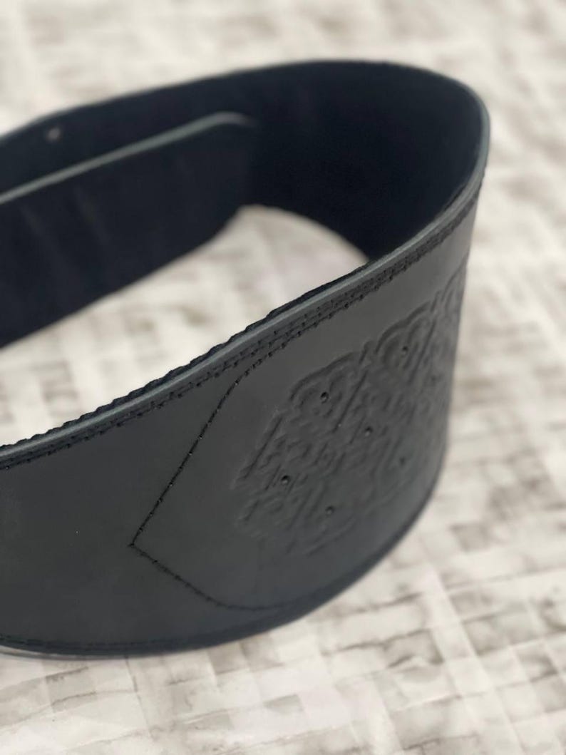May include: A close-up of a black leather belt with an embossed design. The belt is curved, showing the interior lining and stitched edges. The embossed pattern is visible on the front of the belt.
