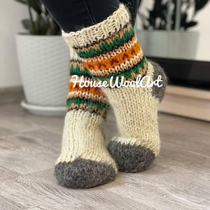 Puede incluir: Un par de calcetines de lana blanca con un patrón de rayas verdes, naranjas y marrones. Los calcetines tienen una puntera y un talón de lana gris.