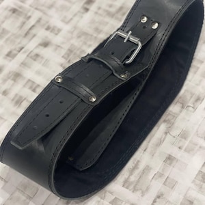 May include: A wide, black leather belt with a silver buckle and rivets. The belt is curved and appears unworn. The leather has a matte finish and is stitched along the edges. The belt is designed to be worn around the waist.
