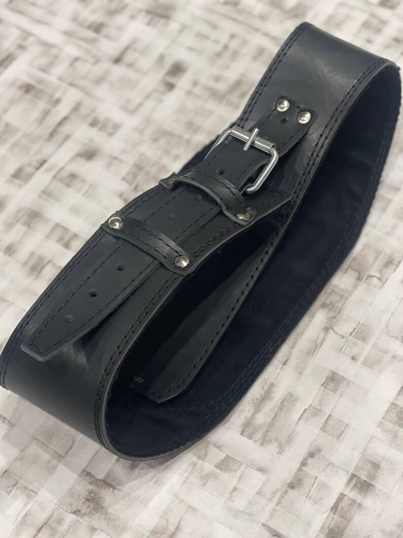 May include: A wide, black leather belt with a silver buckle and rivets. The belt is curved and appears unworn. The leather has a matte finish and is stitched along the edges. The belt is designed to be worn around the waist.