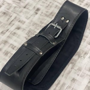 May include: A wide black leather belt featuring a silver buckle and rivets. The belt is made from thick, durable leather. The design includes a buckle and small rivets for added detail, showcasing a robust construction.