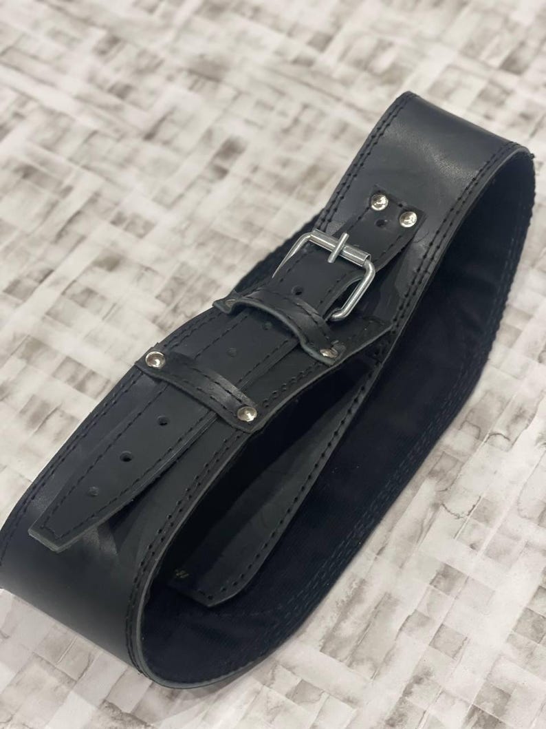 May include: A wide black leather belt featuring a silver buckle and rivets. The belt is made from thick, durable leather. The design includes a buckle and small rivets for added detail, showcasing a robust construction.