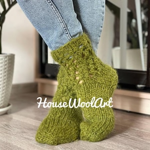 May include: A pair of green knitted socks with a textured pattern. The socks are worn with blue jeans. The text "HouseWoolArt" is visible on the socks. The socks appear to be handmade.