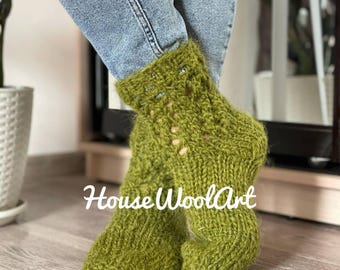 May include: A pair of green knitted socks with a textured pattern. The socks are worn with blue jeans. The text "HouseWoolArt" is visible on the socks. The socks appear to be handmade.