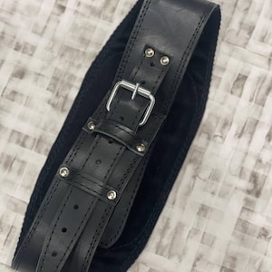 May include: Black leather belt with a silver buckle and rivets. The belt has a wide design with a suede backing. The belt is designed to be worn around the waist.