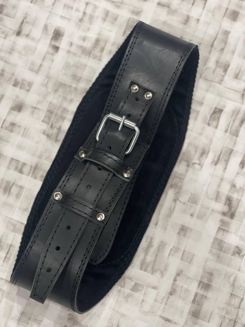May include: Black leather belt with a silver buckle and rivets. The belt has a wide design with a suede backing. The belt is designed to be worn around the waist.