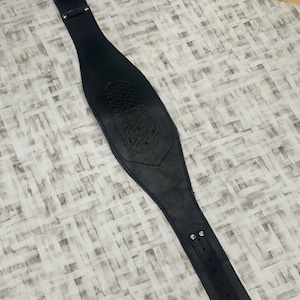 May include: Black leather belt with an embossed central design. The belt features a buckle fastening and is designed for waist wear. The item is displayed on a light-coloured surface. The belt's length is approximately 80-100 cm.