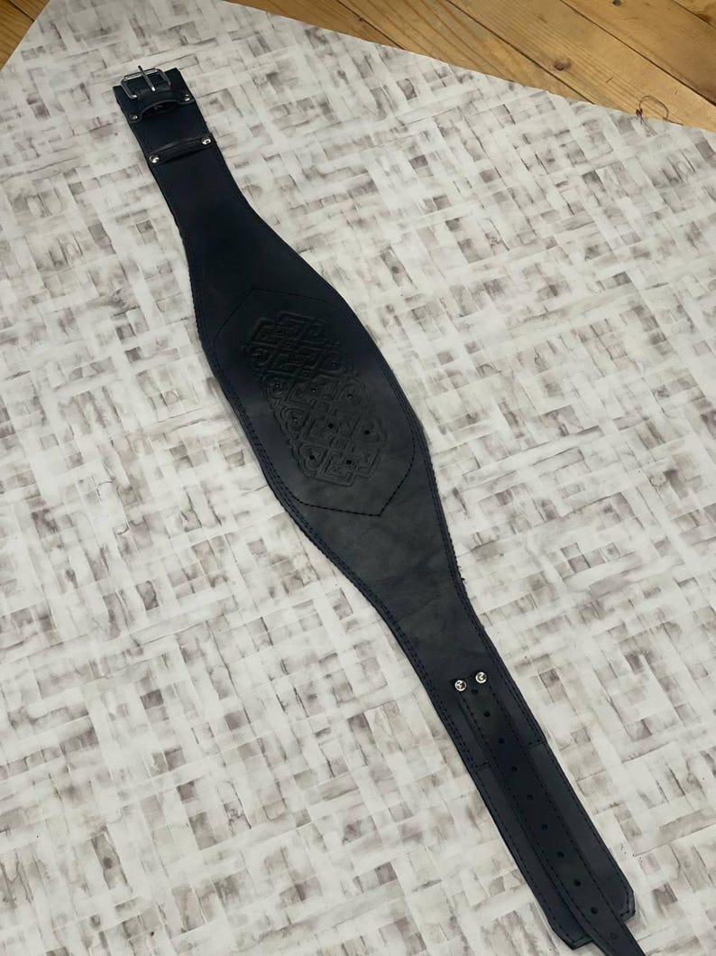 May include: Black leather belt with an embossed central design. The belt features a buckle fastening and is designed for waist wear. The item is displayed on a light-coloured surface. The belt's length is approximately 80-100 cm.