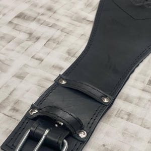 May include: Close-up of a black leather weightlifting belt. The belt features a silver buckle, rivets, and a loop. The leather is stitched along the edges. The belt is designed to provide support during weightlifting exercises.