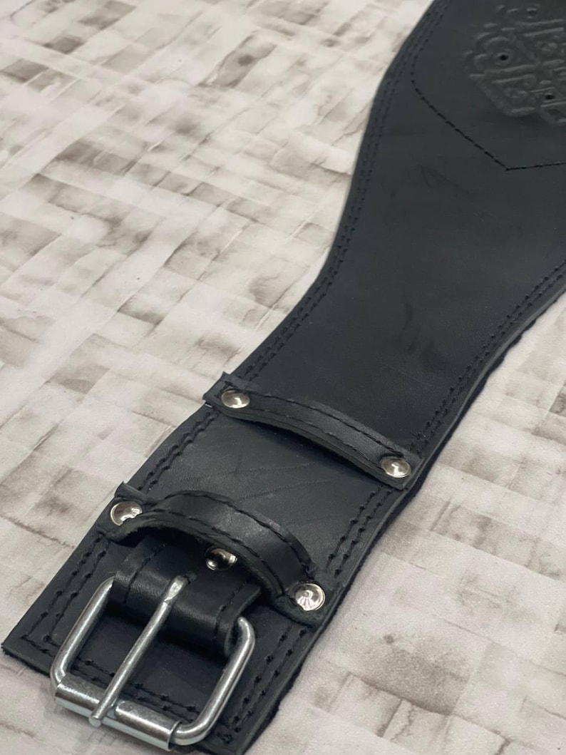 May include: Close-up of a black leather weightlifting belt. The belt features a silver buckle, rivets, and a loop. The leather is stitched along the edges. The belt is designed to provide support during weightlifting exercises.