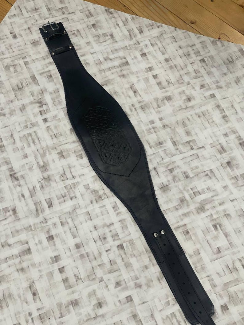 May include: A black leather belt with an embossed design. The belt features a buckle and adjustment holes. The belt is approximately 76 cm long. The design is in the center.