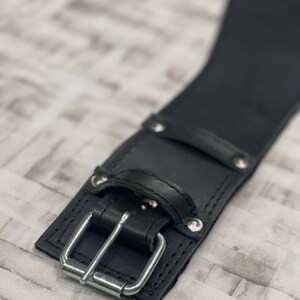 May include: A close-up shot of a black leather belt with a silver buckle. The belt features a rectangular buckle and is secured with rivets. The leather has a smooth appearance, and the design is simple and functional.