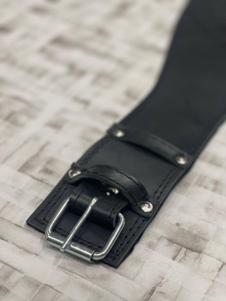May include: A close-up shot of a black leather belt with a silver buckle. The belt features a rectangular buckle and is secured with rivets. The leather has a smooth appearance, and the design is simple and functional.