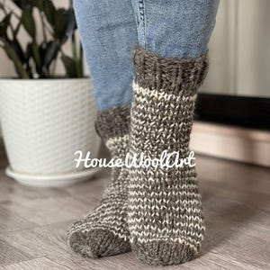 May include: Hand-knitted wool socks in shades of gray and white, with a ribbed cuff. The socks are worn with blue jeans. The text "HouseWoolArt" is visible on the socks. These cozy socks are perfect for cold weather.