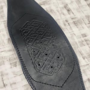 May include: A dark grey leather weightlifting belt with an embossed geometric design. The belt is stitched along the edges and features a decorative pattern in the centre. The design includes small circular cutouts.
