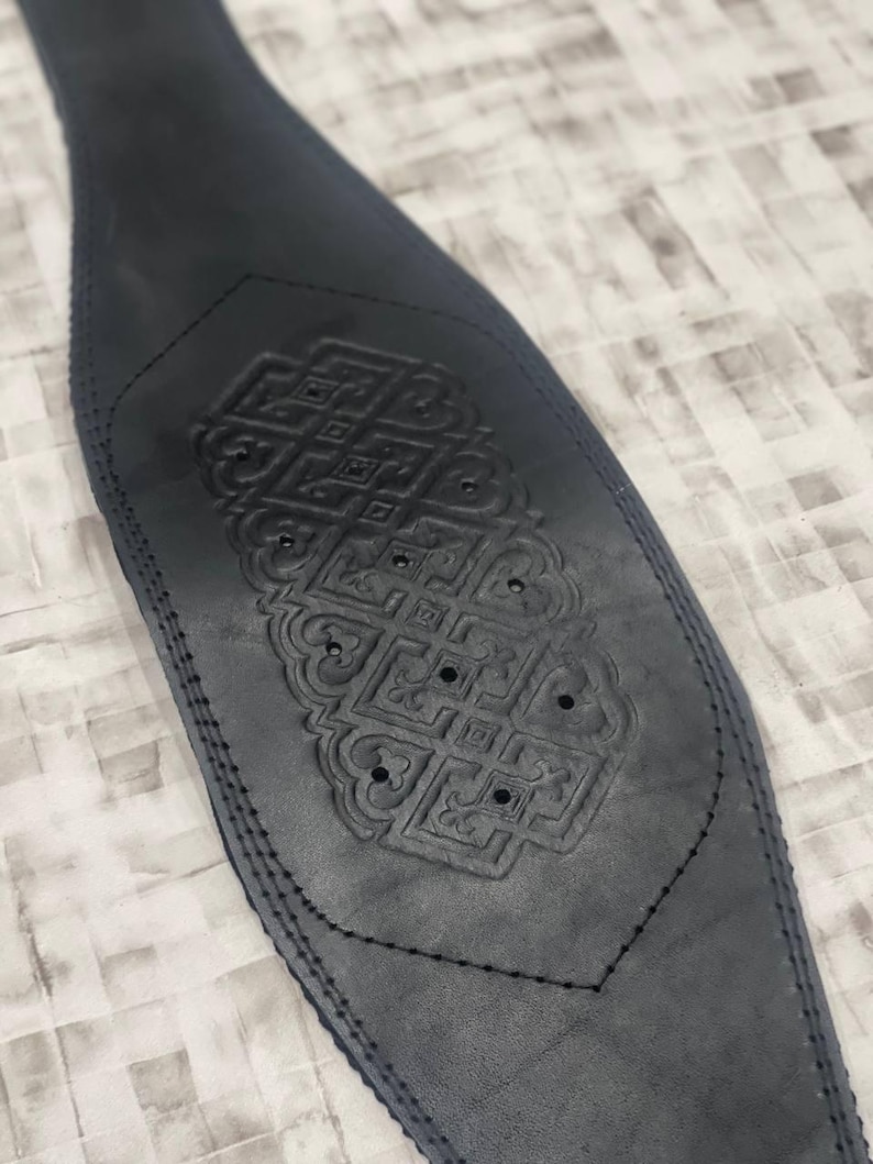 May include: A dark grey leather weightlifting belt with an embossed geometric design. The belt is stitched along the edges and features a decorative pattern in the centre. The design includes small circular cutouts.