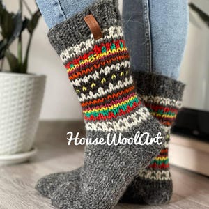 Op de afbeelding: Grijze wollen sokken met een kleurrijk geometrisch patroon in rood, geel, groen en wit. De sokken hebben een geribbelde boord en een leren label. De tekst "House WoolArt" is zichtbaar.