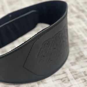 May include: A black weightlifting belt with an embossed pattern. The belt is made of a thick, durable material and has detailed stitching. It is designed to provide support during heavy lifting exercises. The belt's width is approximately 10 cm.