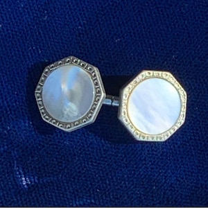 May include: A pair of vintage cufflinks with an octagonal design. Each cufflink features a mother-of-pearl center surrounded by a silver-toned frame with a decorative edge. The cufflinks are connected by a silver bar.