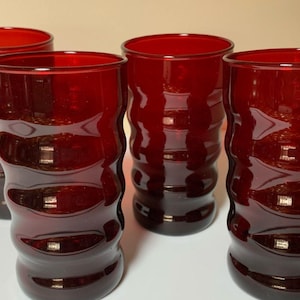 May include: A set of four ruby red drinking glasses. The glasses have a wavy, textured design around the body. The glasses are made of a transparent material, allowing light to pass through. The glasses are suitable for serving beverages.