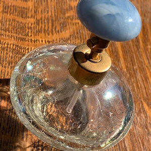 May include: A vintage perfume bottle with a round, cracked glass body and a light blue, marbled top. The bottle has a gold-colored metal neck and a clear glass sprayer. The bottle sits on a wooden surface.