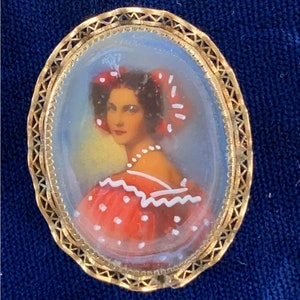 May include: Oval-shaped gold-tone brooch featuring a hand-painted portrait of a woman in a red dress with white polka dots and a matching hair accessory. The portrait is set within a filigree frame, showcasing a vintage aesthetic.