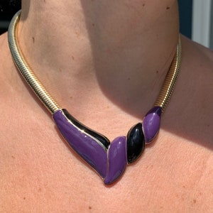 May include: A gold-tone metal collar necklace with a purple and black enamel pendant. The necklace has a flexible, snake-chain design. The pendant features a geometric design with purple and black enamel accents. A stylish statement piece.