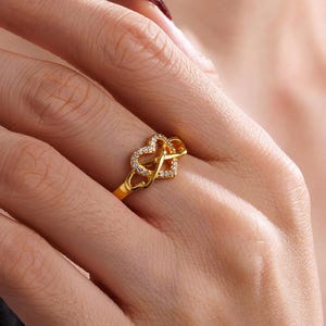 May include: A gold-colored ring featuring two intertwined heart shapes. One heart is encrusted with small, clear gemstones. The ring is worn on a person's finger, showcasing its intricate design and sparkling details. The ring's band is a simple, polished gold.