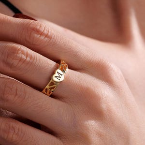 Pode incluir: Um anel dourado com um design em forma de coração e a letra "M" no centro. O anel tem um padrão delicado e vazado. O anel está no dedo de uma pessoa.