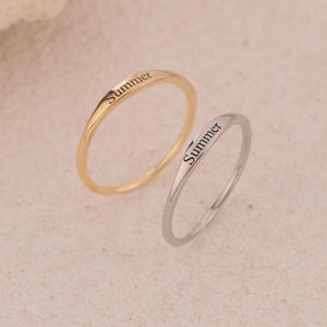 May include: Two slender rings, one gold and one silver, are shown. The word "Summer" is inscribed on each ring in a neat font. The rings are presented on a neutral, textured surface, emphasising their simple, elegant design.