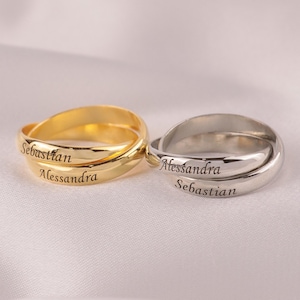 Könnte beinhalten: Zwei personalisierte Ringe, einer goldfarben und einer silberfarben, jeweils mit eingravierten Namen. Der goldfarbene Ring trägt die Inschriften "Sebastian" und "Alessandra". Der silberfarbene Ring trägt "Alessandra" und "Sebastian". Beide Ringe liegen auf einer weißen, seidigen Oberfläche.