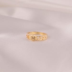 Pode incluir: Um anel dourado com a letra "M" no centro. O anel tem um design em forma de coração com padrões recortados. O anel está sobre uma superfície clara, semelhante a cetim.