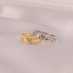 Pode incluir: Dois anéis, um dourado e outro prateado, cada um com um design em forma de coração com a letra "M" gravada no centro. O anel dourado tem uma banda de filigrana, enquanto o anel prateado tem um padrão recortado.