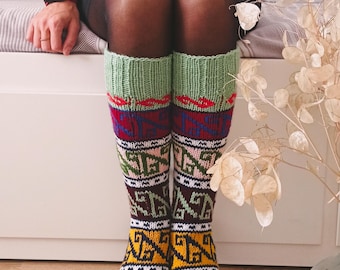 May include: A pair of colorful knitted socks with a geometric pattern. The socks are green, red, yellow, white, and blue. The socks are worn by a person sitting on a couch.