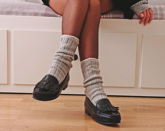 May include: Black leather loafers with tassel detailing. The shoes are paired with grey ribbed socks. The image shows a person sitting on a white bench with floral pillows, wearing a black jacket and a grey sweater.