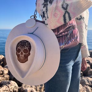 Peut inclure: Un chapeau en feutre blanc avec un motif de crâne de sucre brun. Le chapeau a une large bordure et un ruban turquoise autour de la couronne.